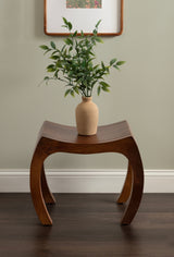 Curved wooden stool with arched legs in warm brown finish, topped with a beige vase holding green leafy branches, set against a pale gray wall with baseboard trim.