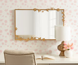 Rectangular wall mirror with a gold metal frame adorned with floral and vine details along the top and sides, reflecting a window and soft pink floral wallpaper.