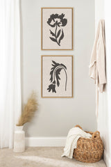 Two framed canvas prints with black botanical silhouettes on textured neutral linen backgrounds, hung vertically on a light gray wall over a wicker basket and vase with dried pampas grass.