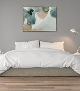 Abstract watercolor-style canvas with muted teal and beige shapes mingling in soft, translucent layers above a white bed with a gray upholstered base, set against a gray wall with a white floor lamp.