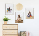 Three framed prints featuring beaded bear cubs in baskets and with piles of laundry, arranged above a light wood dresser and near a woven basket with a pink blanket.