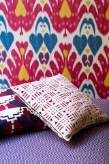 Two decorative pillows with geometric red and navy patterns on a purple textured fabric, set against a colorful wall tapestry featuring repeated heart-like shapes in pink, blue, and yellow.