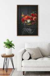 Framed canvas of vibrant red, orange, and white flowers in a brass vase against a dark paneled background, hanging above a white sofa and wooden side table with a green plant.