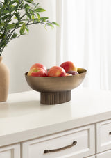 Bronze-colored metal bowl with ribbed cylindrical base holding red apples, placed on white cabinet surface with off-white curtain and leafy branch in background.