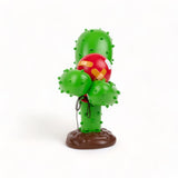 Green cartoon cactus with stubby arms and rounded bumps, featuring a red balloon embedded with orange adhesive bandages, standing on a brown base.