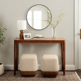 Minimalist wooden console table with tapered legs, topped with a white and brass lamp, a framed photo, and a clear vase with thin branches, paired with two beige fabric ottomans with wooden bases beneath.