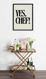 Framed canvas with bold black text 'YES, CHEF!' above a wooden bar cart holding liquor bottles, glassware, a cocktail shaker, and a white tulip bouquet.