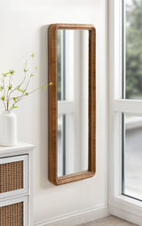 Tall rectangular mirror with a textured, rounded wooden frame mounted on a white wall near a window and a white cabinet with rattan details.