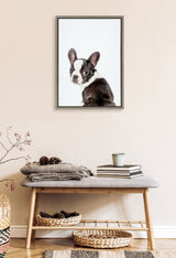 Framed canvas portrait of a black and white French Bulldog with a white background, hanging above a wooden bench with cozy textiles and woven baskets.