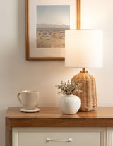 Rattan table lamp with a white drum shade on a wooden console table beside a white vase with dried flowers and a cream ceramic mug under a framed desert landscape photograph.
