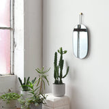 Vertical oval mirror with rounded corners hanging by a thin black strap with brass accent on a white wall, above a white pedestal holding a tall potted cactus and smaller potted plants below.