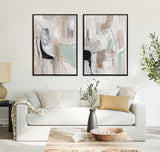 Two framed abstract canvases with soft beige, muted green, black, and brown shapes hang above a white sofa with beige and mustard cushions in a bright living room.