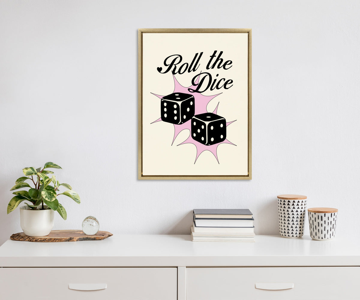 Framed canvas featuring black dice with white dots and the phrase 'Roll the Dice' in black script on a cream background with pink starbursts, hung above a white dresser with plants and patterned containers.