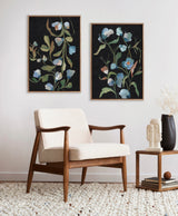 Two framed canvas paintings of delicate blue and white flowers with green leaves on a black background, hanging above a mid-century modern cream armchair with wooden frame.