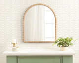 Arched wall mirror with carved wooden frame featuring rounded bead-like accents, mounted above a white mantel with a beige candle holder and a leafy plant in a light pot.