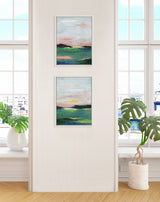 Two framed abstract landscape paintings by Nikita Jariwala in pastel pinks, greens, and blues hung vertically on a beige wall between tall windows with potted plants below.