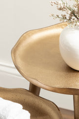 Close-up of a textured gold metal plant stand with a wavy-edged tray holding a white ceramic vase with dried flowers, against a light wall background.