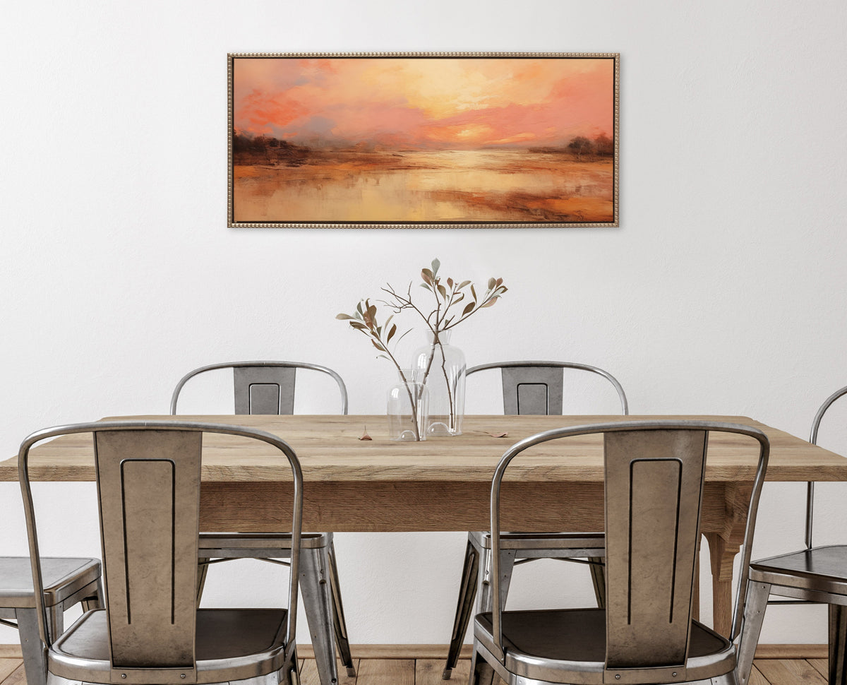Rectangular framed canvas depicting a peach and orange sunset landscape with silhouetted trees, hung above a wooden table and industrial metal chairs in a minimalist room.