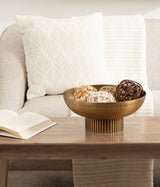 Round ribbed metal bowl with a bronze finish holding decorative woven balls, placed on a wooden coffee table in front of a beige fabric couch with a textured pillow and throw.