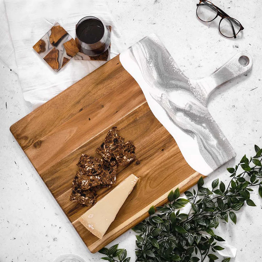 Wooden charcuterie serving tray with cheese and crackers on a white surface with glasses and a plant.