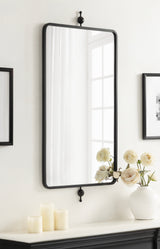 Rectangular wall mirror with a thin black metal frame, mounted with decorative round brackets, reflecting a window and light-colored curtains.