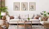 Neutral-toned living room with a beige tufted sofa, three framed botanical prints above, and a round wicker coffee table with plants and books.
