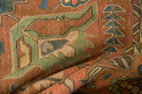 Close-up of a vintage Serapi carpet showcasing earthy brown and muted green geometric patterns with intricate blue leaf motifs on textured fabric folds.