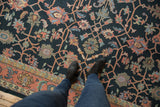 Person standing on a dark floral-patterned vintage carpet with a coral and muted blue border, wooden floor partially visible beneath the carpet edge.