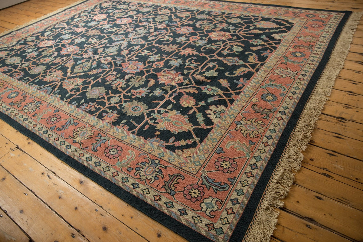 Vintage Serapi Indian Soumac carpet with intricate floral and vine patterns in muted reds, greens, and beiges on a dark background, bordered by a wide reddish-pink band with additional motifs, laid on wooden floor.