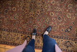 Person wearing black loafers and blue jeans standing on a red and brown Persian-style carpet with intricate floral and medallion patterns.