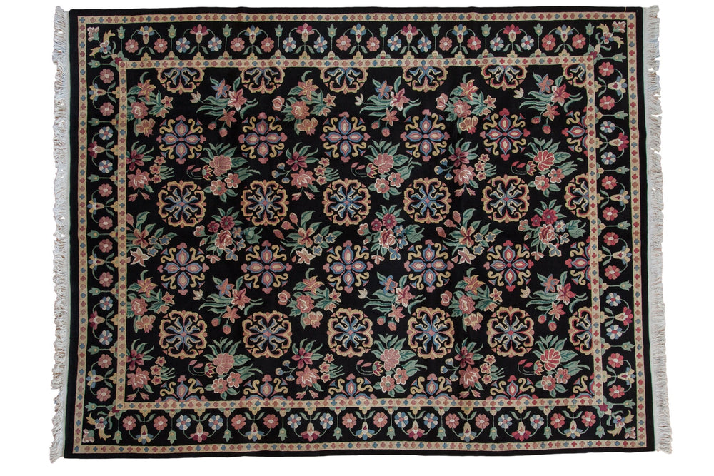 Vintage Indian Arts and Crafts carpet with black background featuring floral and geometric motifs in pink, blue, green, and cream, surrounded by intricate floral borders and cream fringes.