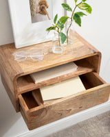 Floating walnut side table with curved edges, an open shelf holding notebooks, and a partially open drawer, topped with two glass vases containing green leafy branches and a photo frame.