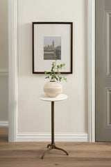 Round side table with white marble top and metal tripod base, holding a textured beige vase with green leafy plant, positioned against a white wall with framed artwork.