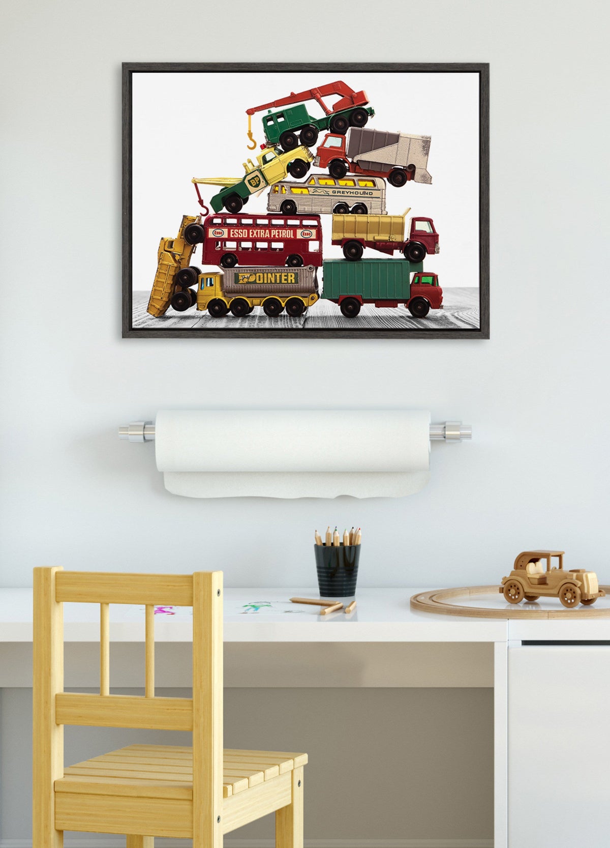 Framed canvas print showing a colorful stack of vintage matchbox work vehicles, including a red crane truck, yellow loader, and a green trailer, mounted on a white wall above a child's desk.