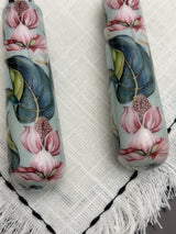 Close-up of floral-themed cake knife and server handles decorated with pink flowers and green-blue leaves on a light background, resting on white textured fabric with tassels.
