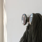 Two metallic, round Sequence coat hooks with a polished mercury finish mounted on a white wall, one holding a draped dark green fabric.