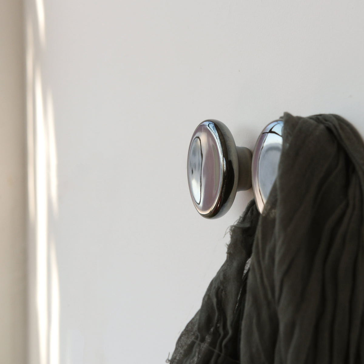 Two metallic, round Sequence coat hooks with a polished mercury finish mounted on a white wall, one holding a draped dark green fabric.