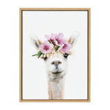 Close-up portrait of a light-colored alpaca wearing a crown of pink flowers and green leaves against a white background in a simple wooden frame.
