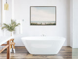 White freestanding bathtub centered on wood floor planks beneath framed beach scene with pale sky, green grasses, and sandy path, flanked by wooden stool and glass pendant light.