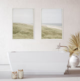 Minimalist bathroom featuring a modern white freestanding tub beneath two framed coastal landscape canvases depicting grassy dunes and a calm sea under soft gray skies.