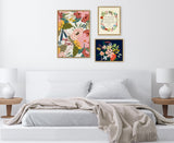 Three framed artworks above a white bed: large pink and yellow floral print, small floral print with text on cream background, and small dark floral print with pink, orange, and yellow flowers.