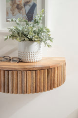 Close-up of a wooden floating side table with vertical ribbed cylindrical slats under a smooth round top, featuring a white textured ceramic pot with green foliage and eyeglasses nearby.