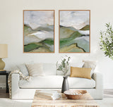 Two framed abstract landscape paintings above a white sofa, featuring muted greens, whites, and browns with soft, brushstroke patterns suggesting hills and sky.