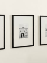 Black photo frame with small bead detailing along the edges, displaying a black-and-white photo with a white mat, mounted on a beige wall.