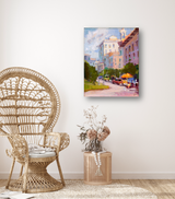 Impressionist-style canvas painting of a city street with pastel buildings, greenery, and a bright orange umbrella under sunlight, hanging on a cream wall above a wicker table and chair.