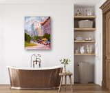 A copper freestanding bathtub with vintage-style faucet in a bright bathroom featuring wooden flooring, a small wooden stool with a vase of flowers, and an impressionist cityscape painting on the wall.