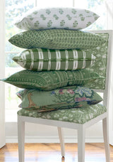 Stack of six green and white patterned pillows with floral and geometric designs on a white chair with a green floral cushion, in front of a window.