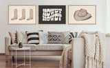 Three framed minimalist prints above a beige couch with patterned pillows and a chunky knit throw, featuring two cowboy boots, 'HOWDY HOWDY' text, and a cowboy hat in muted tones.