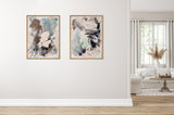 Two abstract framed canvases with brushstrokes in blues, grays, blacks, and whites hanging side by side on a white wall above light wood flooring in a bright living room.