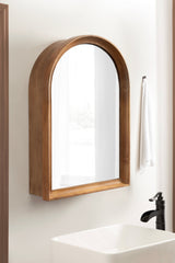 Arched wall mirror with a smooth medium wood frame mounted above a white rectangular sink and black faucet in a minimalist bathroom.
